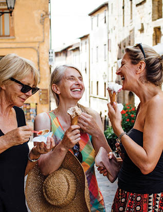 Three mature female friends standing eating Italian ice-creams while in a street in Tuscany during summer. They are smiling and facing each other and enjoying their holiday.