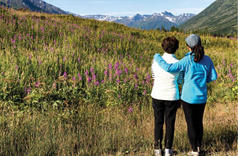 Mother and daughter, backs to camera, looking at wild flowers in field with mountains and sky in background. Selective focus on people.